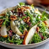 Fresh frisée lettuce topped with sliced pears, crumbled blue cheese, and crispy prosciutto, drizzled with a zesty vinaigrette in a white ceramic bowl.