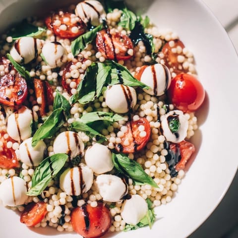 Caprese Couscous Salad is garnished with fresh basil leaves and a generous drizzle of glossy balsamic glaze on a rustic wooden table.  